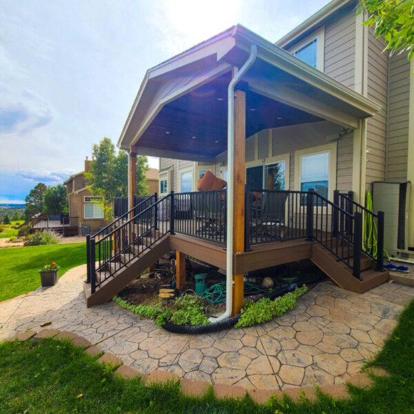 Covered wooden patio attached to a house with landscaped garden around.